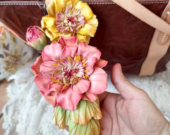 May include: A leather flower charm with a pink and yellow flower design. The charm is attached to a brown leather bag.