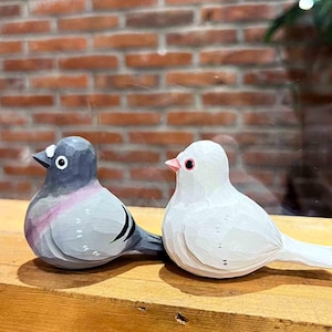 以下が含まれることがあります： 木製の鳥の置物2つ。1つはグレーでピンクのアクセント、もう1つは白。木製の台の上に置かれています。鳥は細かな彫刻とペイントが施されています。
