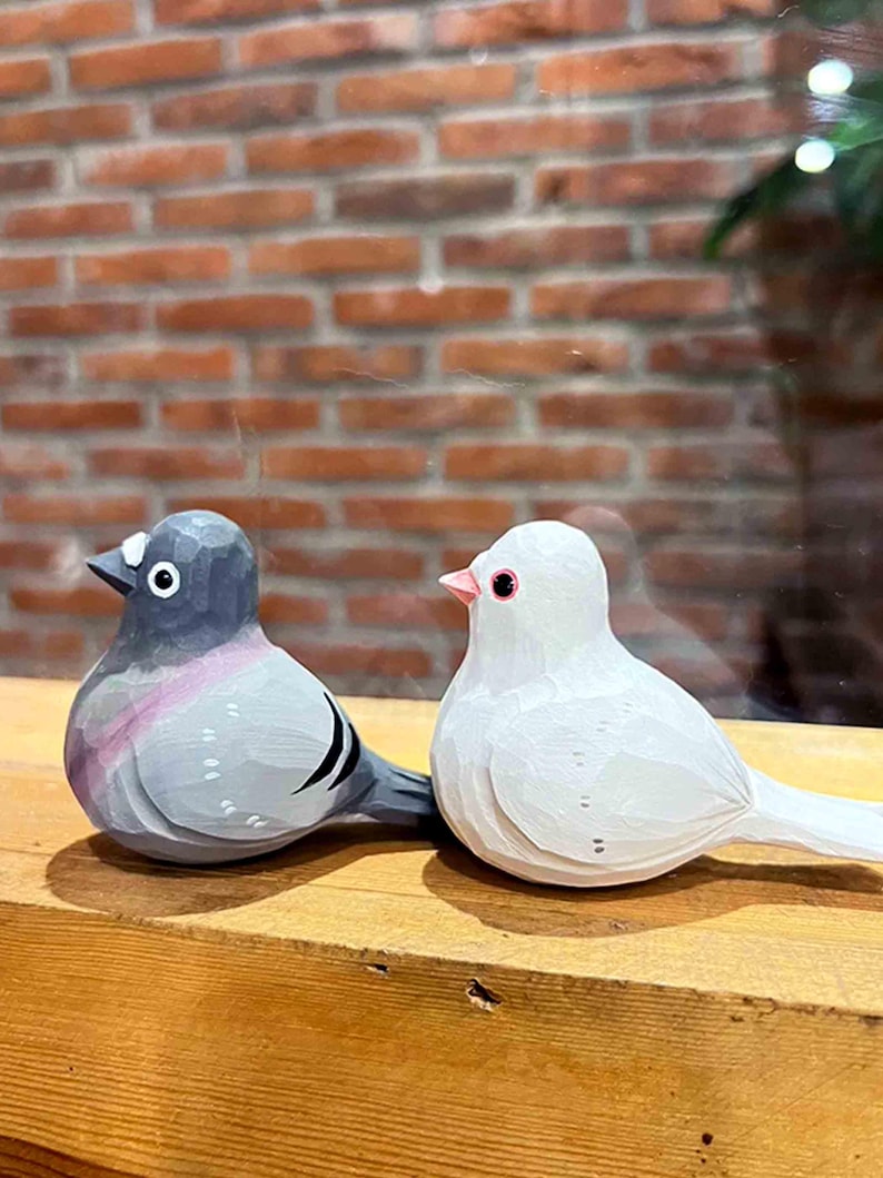 以下が含まれることがあります： 木製の鳥の置物2つ。1つはグレーでピンクのアクセント、もう1つは白。木製の台の上に置かれています。鳥は細かな彫刻とペイントが施されています。