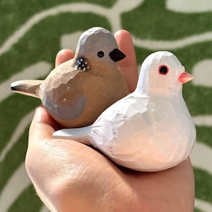 以下が含まれることがあります： 木製の鳥の置物2つ。1つは茶色で灰色のアクセント、もう1つは白でピンクのくちばし。手のひらに乗せられている。