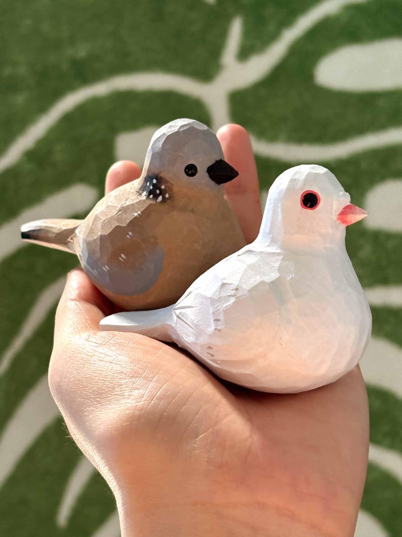 以下が含まれることがあります： 木製の鳥の置物2つ。1つは茶色で灰色のアクセント、もう1つは白でピンクのくちばし。手のひらに乗せられている。