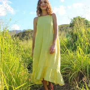 May include: A woman wearing a yellow gingham print dress with a ruffled hem stands in a field of tall green grass. The dress has a square neckline and thin straps. The sky is blue with some clouds.