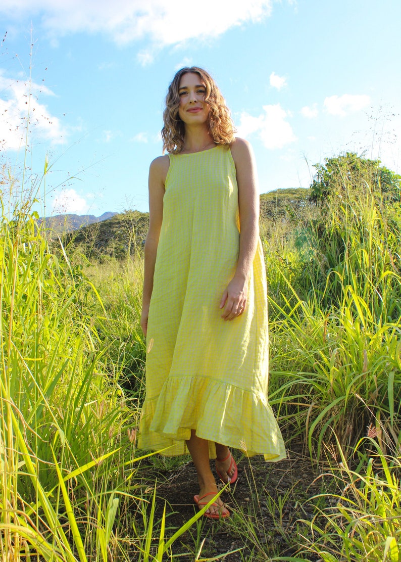 May include: A woman wearing a yellow gingham print dress with a ruffled hem stands in a field of tall green grass. The dress has a square neckline and thin straps. The sky is blue with some clouds.