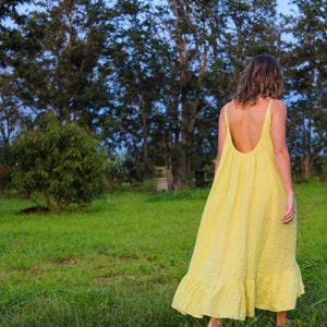 May include: A yellow sundress with a low back and spaghetti straps. The dress has a ruffled hem and is worn outdoors in a grassy field. Trees and a blue sky are in the background.