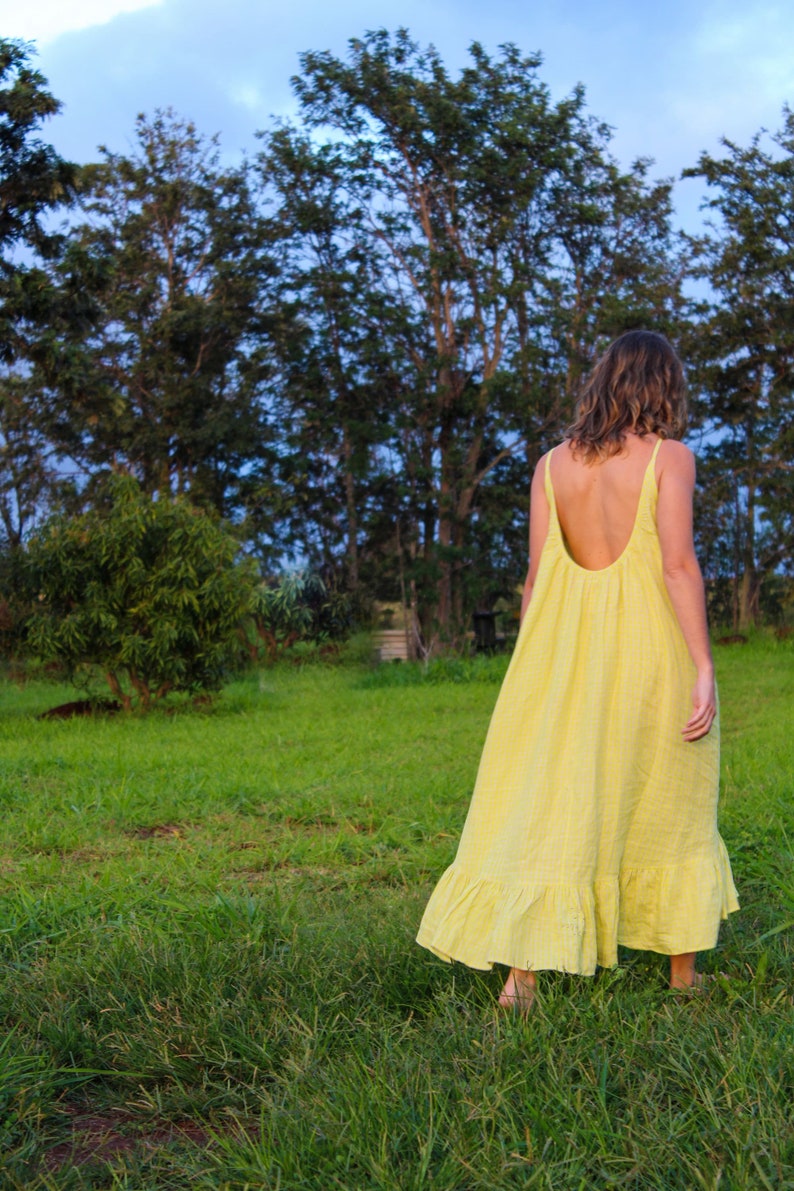 May include: A yellow sundress with a low back and spaghetti straps. The dress has a ruffled hem and is worn outdoors in a grassy field. Trees and a blue sky are in the background.