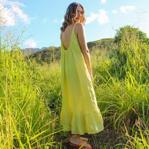 May include: A woman wearing a yellow, sleeveless maxi dress with a low back, standing in a field of tall green grass. The dress has a ruffled hem and is paired with orange sandals. The sky is blue with fluffy white clouds.