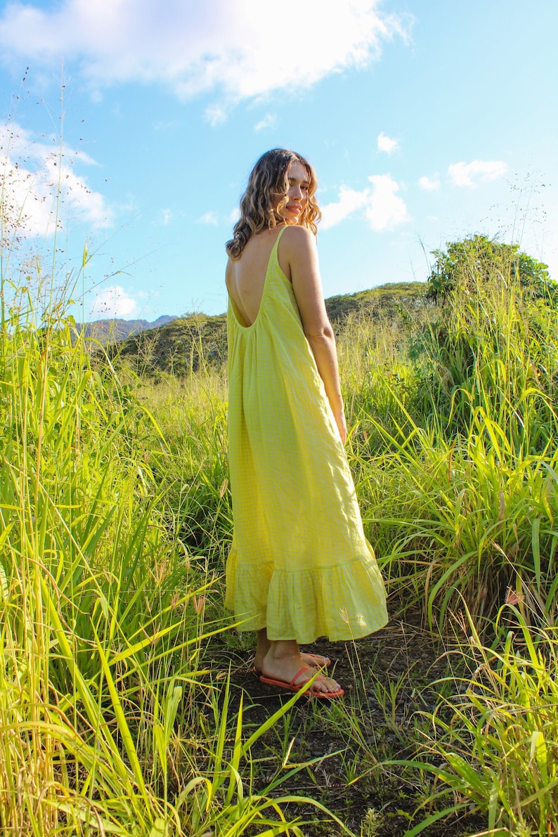 May include: A woman wearing a yellow, sleeveless maxi dress with a low back, standing in a field of tall green grass. The dress has a ruffled hem and is paired with orange sandals. The sky is blue with fluffy white clouds.