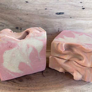 May include: Two bars of handmade soap with a pink and white swirl pattern. The soap is on a wooden surface.