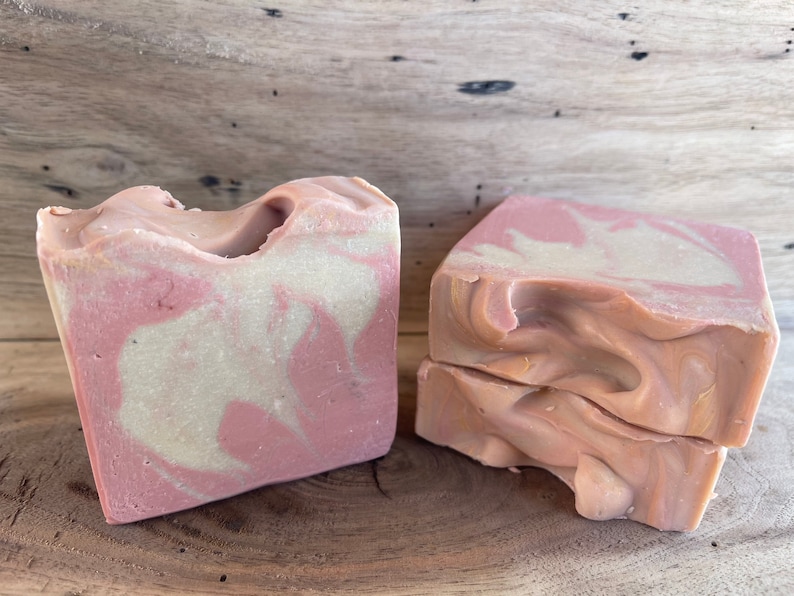 May include: Two bars of handmade soap with a pink and white swirl pattern. The soap is on a wooden surface.