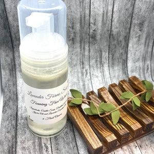 May include: A clear plastic bottle with a white label that reads "Lavender Farms Soap Foaming Face Wash" and lists the ingredients as "Castle Soap, Distilled Water, Jojoba Oil and Lavender Essential Oil". The bottle is sitting on a wooden surface with a green vine.
