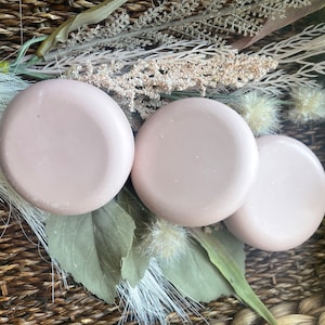 May include: Three round, light pink soap bars in a wicker basket with dried flowers and greenery.