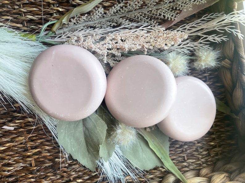 May include: Three round, light pink soap bars in a wicker basket with dried flowers and greenery.