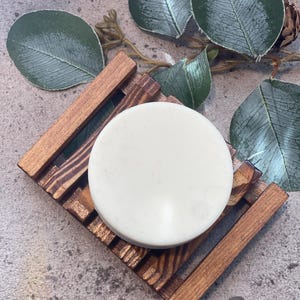 Puede incluir: Una pastilla de jabón redonda y blanquecina descansa sobre un jabonera de madera con diseño de listones. La jabonera es de madera teñida. Hojas verdes y una piña en el fondo sugieren un tema natural.