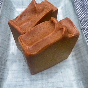 May include: Two brown bars of soap on a light blue ceramic dish. The soap is handmade and has a natural, earthy look.