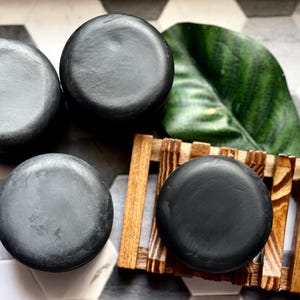 Puede incluir: Cuatro jabones redondos de color gris oscuro. Una pastilla de jabón descansa sobre un pequeño jabonero de madera. Una gran hoja verde es visible en el fondo. Las pastillas de jabón tienen un acabado liso y mate. La disposición sugiere un tema de spa o cuidado personal.