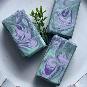 May include: Three rectangular bars of handmade soap with a marbled design. The soap features swirls of light green and lavender colors. A small sprig of green leaves is placed between the bars. The soaps are arranged on a white, textured surface.