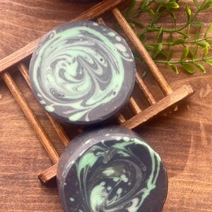 Puede incluir: Dos barras redondas de jab&oacute;n con un dise&ntilde;o de espiral. El jab&oacute;n es negro con remolinos verdes. El jab&oacute;n est&aacute; sobre un jabonera de madera.
