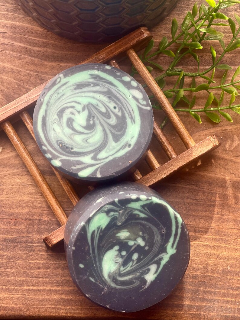 Puede incluir: Dos barras redondas de jab&oacute;n con un dise&ntilde;o de espiral. El jab&oacute;n es negro con remolinos verdes. El jab&oacute;n est&aacute; sobre un jabonera de madera.