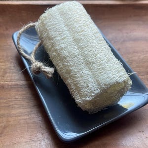 May include: A natural loofah sponge with a jute rope loop rests on a rectangular, dark gray ceramic soap dish. The loofah is light beige and has a textured, porous surface. The dish sits on a wooden surface.