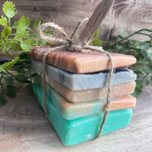May include: A stack of four handmade soap bars wrapped in twine. The bars are in shades of brown, blue, green, and beige.