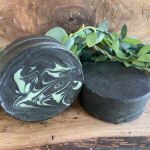Puede incluir: Dos barras redondas de jab&oacute;n negro y verde. El jab&oacute;n de la izquierda tiene un patr&oacute;n de remolino de verde y negro. El jab&oacute;n de la derecha es de color negro s&oacute;lido.