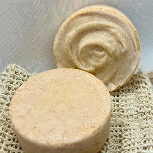 May include: Two round, light brown soap bars on a white mesh surface. The soap bars have a swirl pattern and a rough texture.