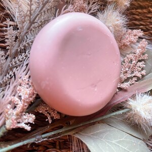 May include: A round, pale pink soap bar with a smooth surface. The soap is sitting on a woven basket with dried flowers.