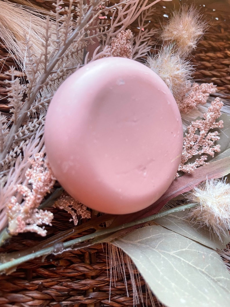 May include: A round, pale pink soap bar with a smooth surface. The soap is sitting on a woven basket with dried flowers.