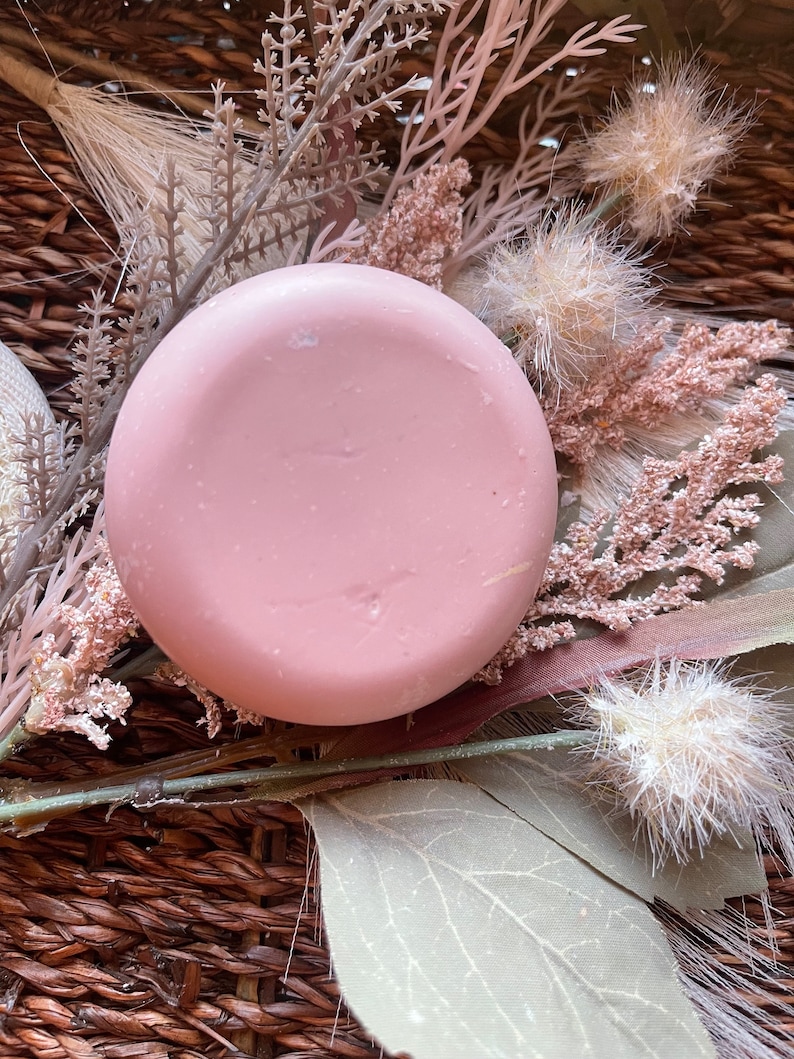 May include: A round, light pink soap bar with a smooth surface, surrounded by dried flowers and leaves.