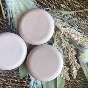 May include: Three round, light pink, solid color soap bars on a woven brown basket surface with dried flowers and grasses.