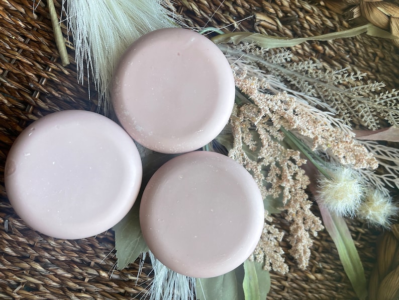 May include: Three round, light pink, solid color soap bars on a woven brown basket surface with dried flowers and grasses.