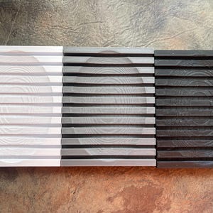 Puede incluir: Tres posavasos rectangulares dispuestos uno al lado del otro, con un diseño de listones horizontales y una hendidura circular en el centro. Los posavasos son de color blanco, gris y negro, cada uno con una textura de veta de madera visible.