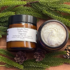 May include: Two jars of Lavender Farms Tallow Butter Peppermint, one open, revealing a swirl of white cream. The closed jar has a label with product information. The jars are surrounded by evergreen branches, pinecones, and red berries.