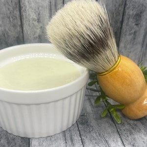May include: A white ceramic bowl filled with a light green shaving cream and a wooden shaving brush with a gold band and white bristles.