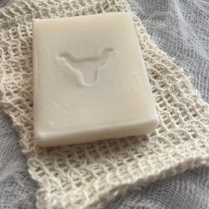 May include: A rectangular off-white bar of soap featuring an embossed bull head logo on its surface, resting on a beige woven mesh drawstring pouch with a small brown wooden bead at the top closure, showcasing a simple and functional design.