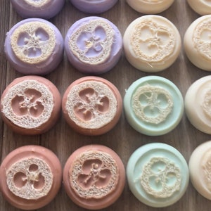 May include: An assortment of round soaps in various pastel colors, including lavender, peach, and mint green. Each soap features a natural loofah embedded in the center, creating a textured surface. The soaps are arranged in neat rows.