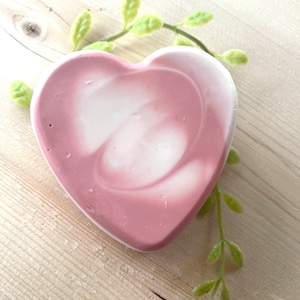 May include: A heart-shaped soap bar with a swirl design in shades of pink and white. The soap is sitting on a wooden surface with green leaves.
