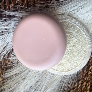May include: A round, light pink soap bar on top of a white loofah sponge. The soap and sponge are on a woven brown background.
