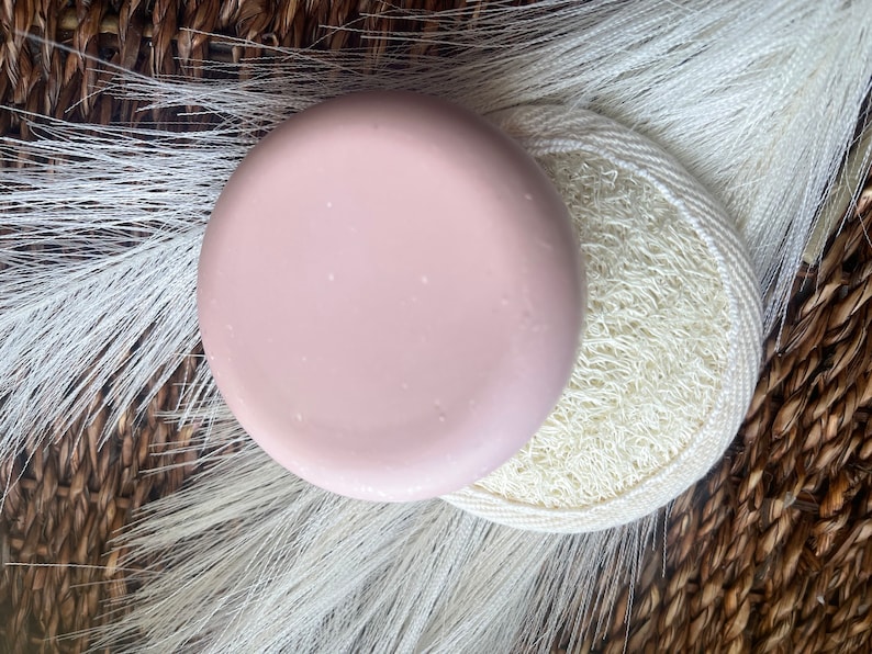May include: A round, light pink soap bar on top of a white loofah sponge. The soap and sponge are on a woven brown background.
