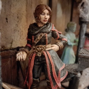 Op de afbeelding: Een geschilderd miniatuurbeeldje van een vrouw die een donkere kapmantel met rode accenten draagt. Ze heeft lang bruin haar en houdt een dolk in haar rechterhand.