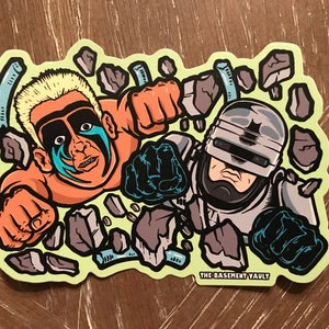 May include: A sticker featuring a cartoon illustration of a wrestler with blue face paint and a cyborg police officer, both punching through a wall of broken concrete. The sticker has a yellow background and the text "THE BASEMENT VAULT" at the bottom.