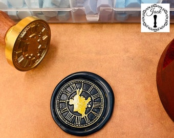 May include: A black wax seal with a gold clock face design. The clock face features a stylised image of a rabbit with a key in its hand. The seal is on a brown surface.