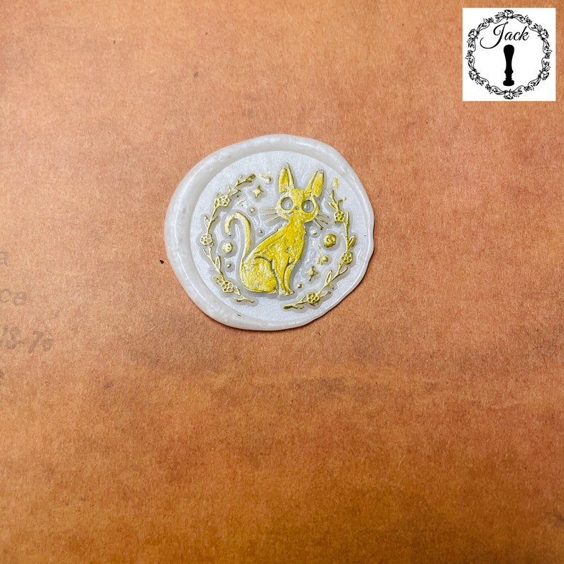 May include: A white wax seal with a gold design of a cat with stars and flowers around it.