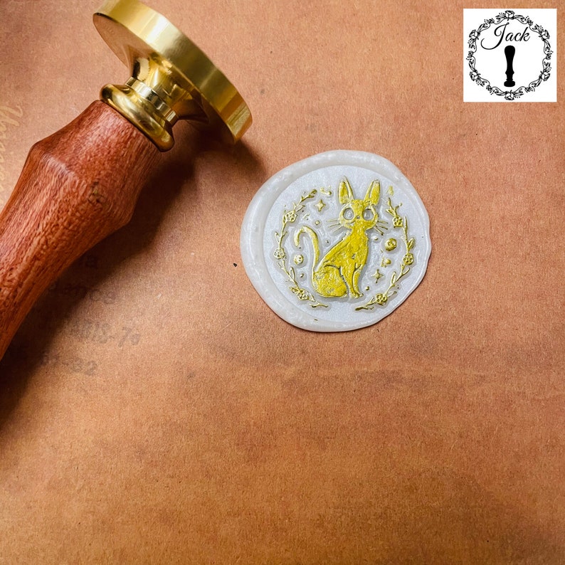 May include: A white wax seal with a gold design of a cat sitting in a wreath of flowers. The cat has a golden star above its head.