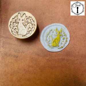 May include: A brass stamp featuring a design of a cat sitting with stars and flowers around it. The stamp is used to create wax seals.
