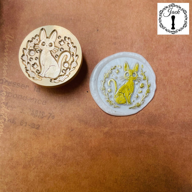 May include: A brass stamp featuring a design of a cat sitting with stars and flowers around it. The stamp is used to create wax seals.