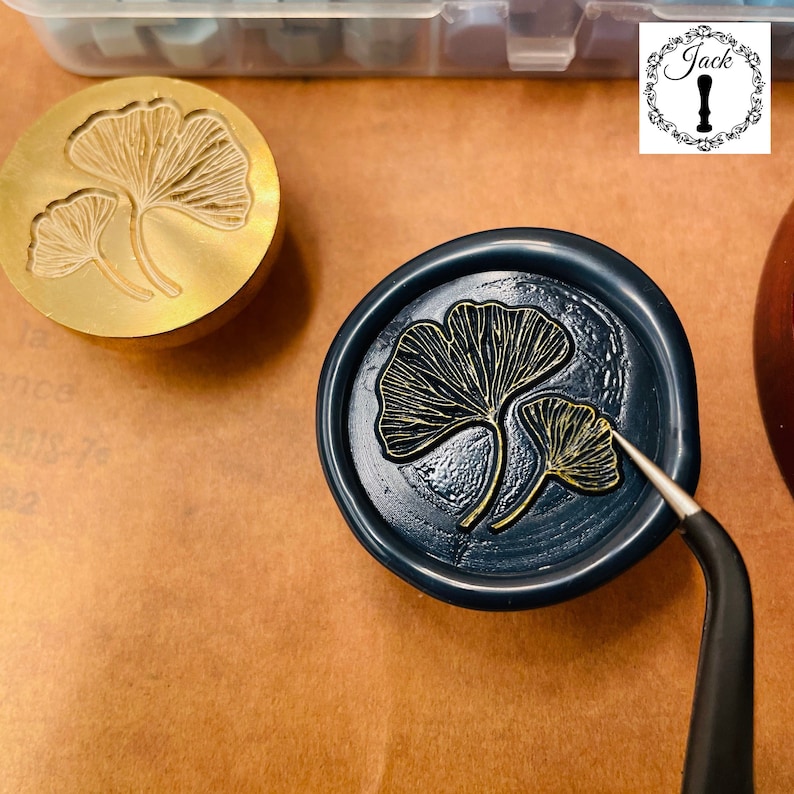 May include: A gold metal stamp featuring a design of two ginkgo leaves. The stamp is being used to create a wax seal with a blue background and gold ginkgo leaves.
