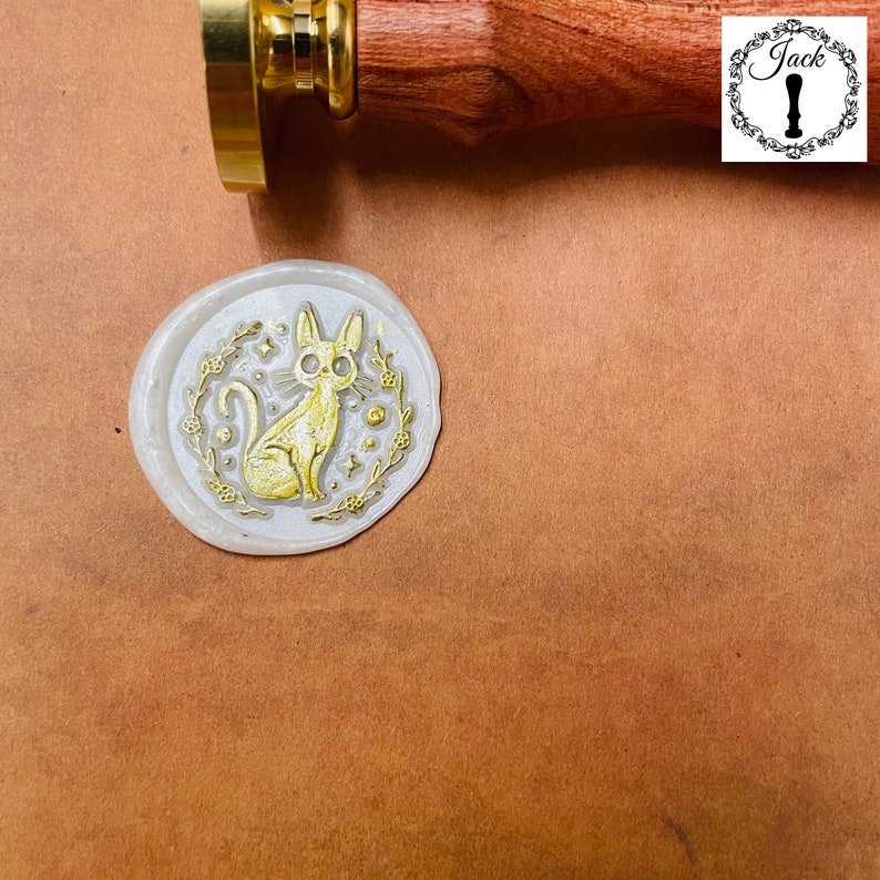 May include: A white wax seal with a gold design of a cat with stars and leaves. The seal is on a brown surface.