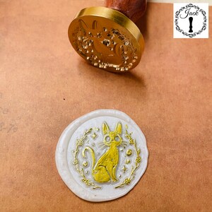 May include: A white wax seal with a gold design of a cat with a floral wreath around it.