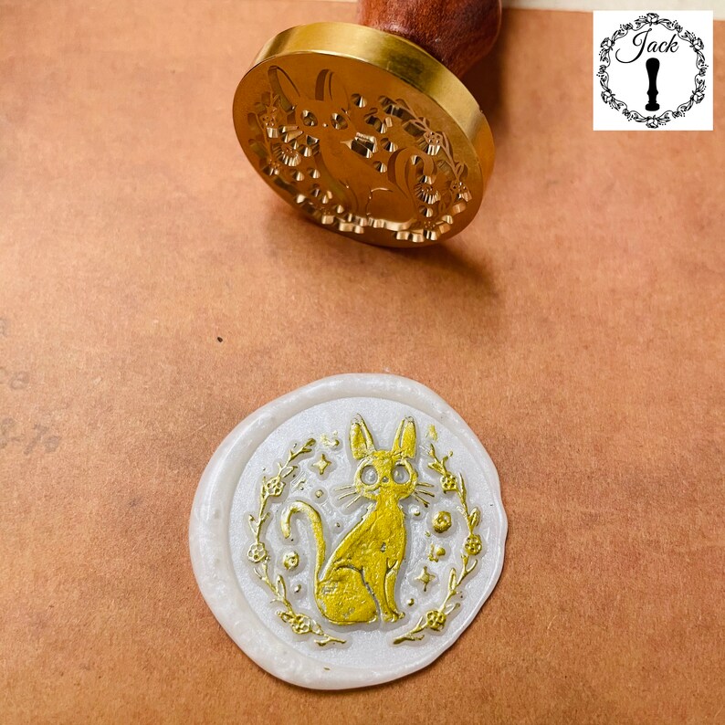 May include: A white wax seal with a gold design of a cat with a floral wreath around it.